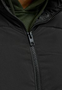 Musta toppatakki vetoketjulla, jossa sileä pinta ja tummanvihreä huppari näkyvissä kaula-aukon kohdalta.