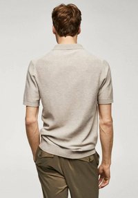 Hombre con cabello corto castaño, vistiendo una camiseta de polo de punto de manga corta color beige y pantalones verdes oliva, de espaldas contra un fondo sencillo.