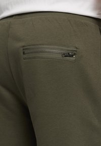 Gros plan sur la poche arrière d'un pantalon vert olive avec fermeture éclair horizontale et taille élastique, porté avec un haut blanc.