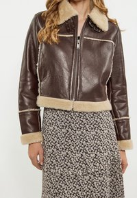 Brązowa krótka skórzana kurtka z beżowym kołnierzem i mankietami z baranka; posiada przedni zamek błyskawiczny i kontrastowe przeszycia, noszona na kwiecistej spódnicy.