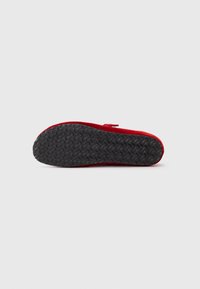 Zapato rojo con suela de goma negra texturizada, que cuenta con un material superior suave y una correa para un ajuste seguro, visto desde la parte inferior.