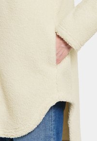 Cremefarvet fleece top med en ujævn kant og sidelommer, der har en blød tekstur og lange ærmer. Jeans ses i bunden.