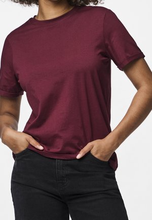 Person iført en ensfarvet burgunder kortærmet t-shirt, let stoppet ind i sorte bukser med hænderne i lommerne mod en hvid baggrund.