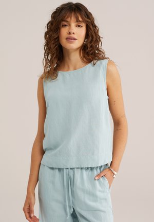 Naisel lokkidega pruunide lokkis juustega, kandes helesinist varrukateta toppi ja sobivaid lahtisi pükse, seisab ühe käega taskus.