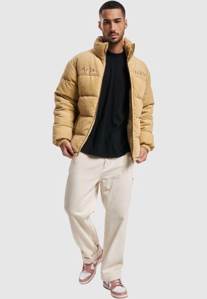 Giacca puffer in corduroy color tan con design trapuntato, colletto alto e ricamo "KARL". Abbinata a una camicia nera e pantaloni cargo color crema.
