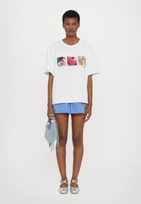 Hvit oversized t-skjorte med fire bilder på fronten, kombinert med blåstripete shorts. Holder en grå fransete håndveske, iført sølvfargede sko.