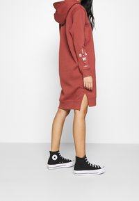 Červené šaty s kapucí, s bočními rozparky, žebrovanými manžetami a potiskem na jednom rukávu. Kombinované s černými kotníkovými teniskami.