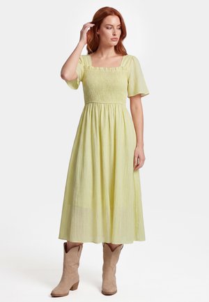 Frau mit roten Haaren trägt ein hellgelbes Midikleid mit flatternden Ärmeln und beigen Stiefeln bis zur Mitte der Wade, steht mit einer Hand in der Nähe ihrer Haare.