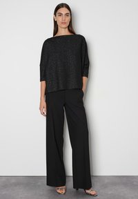 Schwarzes, oversized Oberteil mit glitzernder Textur und Bootsausschnitt, kombiniert mit schwarzen weiten Stoffhosen. Model trägt silberne Riemchensandalen.