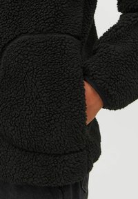 Giacca in pile nera realizzata con un materiale texturizzato e soffice. Presenta una tasca laterale e una vestibilità aderente intorno al polso, che ne migliora il calore.