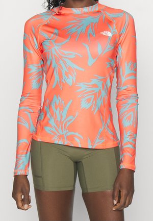 Vrouw draagt een feloranje longsleeve met blauwe bloemmotieven en olijfgroene aansluitende short, staand tegen een effen achtergrond.