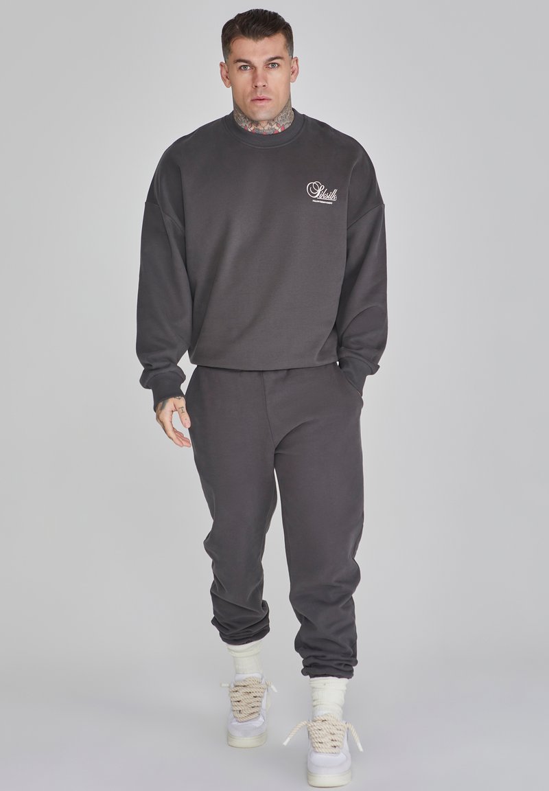 Sudadera gris oscuro y joggers a juego hechos de material suave. Presenta un cuello redondo, corte holgado y puños acanalados, con un pequeño detalle de logo.