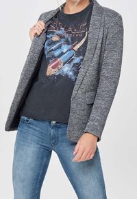 Blazer gris texturizado con cuello chal, combinado con una camiseta gráfica negra y jeans azules, cuenta con dos bolsillos frontales.