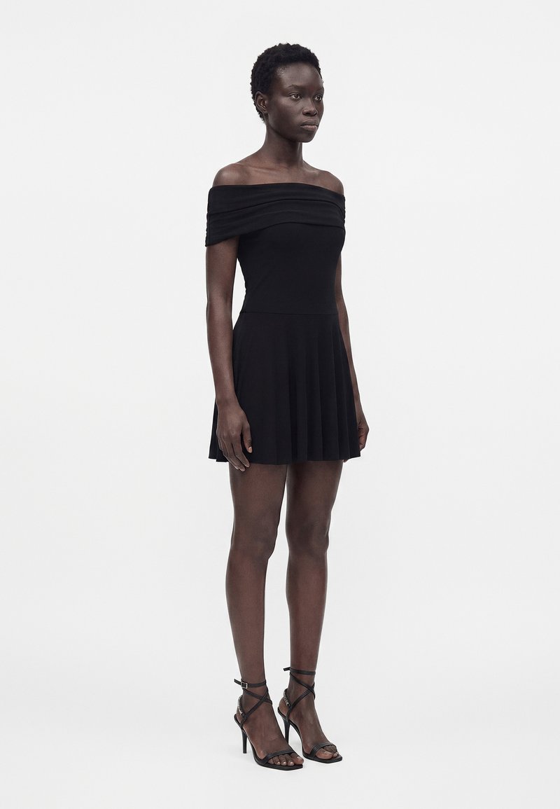 Femme debout, portant une robe courte noire épaules dénudées et des sandales noires à talons hauts, sur un fond clair uni.