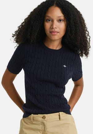 Pull en maille côtelée bleu marine avec des manches courtes et un col rond, présentant un motif en cable et un petit logo au niveau de la poitrine.