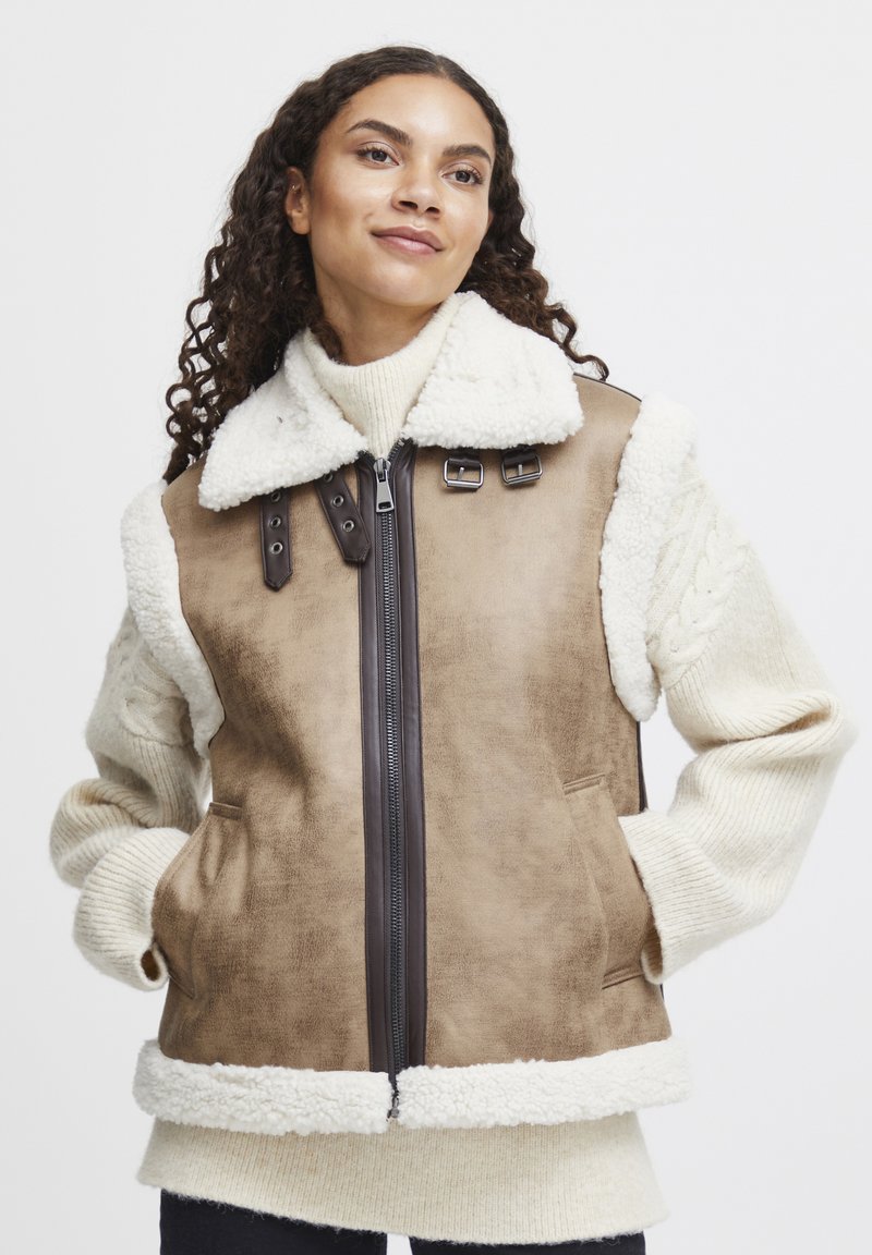 Bruine nepleer vest met crèmekleurige schapenvacht kraag en accenten. Zip-closure, zijzakken en gemêleerde gebreide mouwen in ivoor.