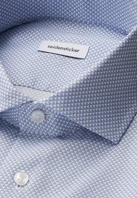 Camicia azzurra chiara con un piccolo motivo triangolare blu scuro, bottoni bianchi e un'etichetta "seidensticker" all'interno del colletto.