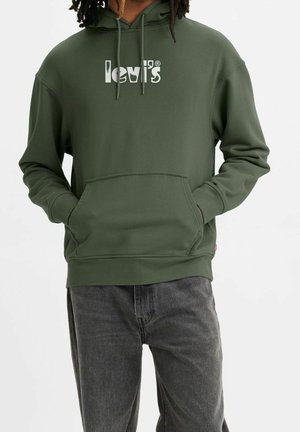 Personne portant un sweat à capuche Levi's vert olive avec poche kangourou et jean noir, les mains dans la poche, sur un fond blanc uni.