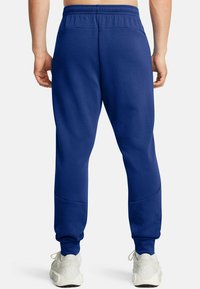 Donkerblauwe sweats met een elastische tailleband, taps toelopende benen en kniestukken, gecombineerd met lichtgrijze sportschoenen.
