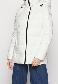 Vit puffjacka med dragkedja, utrustad med huva och elastiska snoddar. Tillverkad av ett slätt, vadderat material med sidofickor.