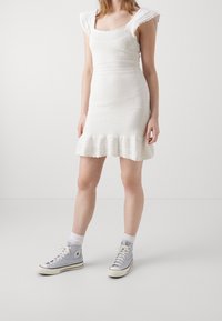 Robe blanche en tricot avec un col carré, des manches courtes à volants et un ourlet évasé. Associée à des baskets montantes bleu clair.