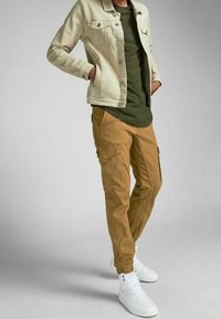 Beige denimjacka över en olivgrön T-shirt, tillsammans med bruna cargo-byxor med flera fickor, och vita högklackade sneakers.