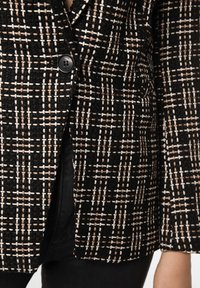 Blazer à carreaux noirs et beiges en tissu texturé, avec un unique bouton noir, des revers définis et des bords soigneusement finis.