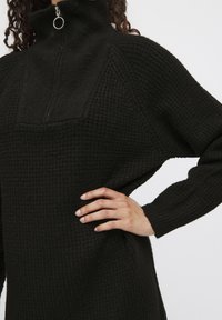 Čierny, textúrovaný pulover s vysokým golierom a dizajnom na polovičný zips. Obsahuje dlhé rukávy a voľný strih, s ribbovanými akcentmi na pleciach.