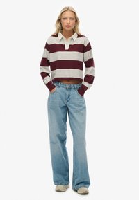 Kvinde står med hænderne i lommerne iført en cropped maroon og grå stribet langærmet skjorte, løse lyseblå jeans og hvide sneakers.