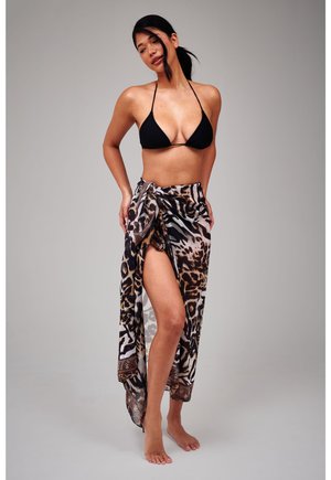 Woman standing barefoot wearing a black bikini top and a leopard-print sarong tied at the waist against a gray background.