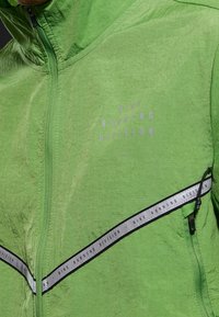 Veste verte avec une finition texturée. Dispose d'une fermeture éclair à l'avant, d'accents réfléchissants et d'un logo imprimé en gris clair sur la poitrine.