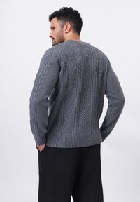 Pull en tricot gris avec un motif de câbles texturé, des poignets et un ourlet côtelés. Le dos présente des motifs diagonaux et en losanges.