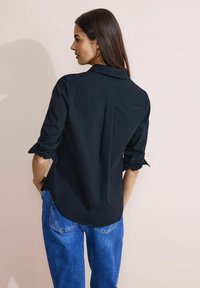 Donkerblauwe, langemmouwshirt van gladde stof; de achterkant heeft een plooi-design. Gecombineerd met blauwe jeans die van opzij zijn weergegeven.