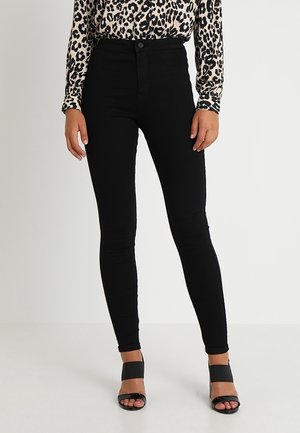 Femme portant un jean skinny noir taille haute, un top à manches longues imprimé léopard et des talons ouverts noirs, debout devant un fond uni.