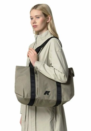 Giovane donna con giacca beige che porta una grande borsa a tracolla beige con spalline nere sulla spalla, su sfondo bianco.