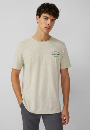 Lichtbeige T-shirt met korte mouwen in een zachte textuur, met een groen grafisch logo op de linkerborst en een ronde hals.