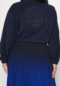 En person som bär en marinblå Adidas fleecejacka med texten "ATHLETIC LEAGUE" på ryggen och en veckad kjol med en svart till blå gradient.