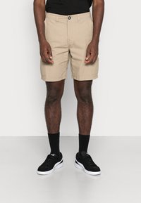 Homme portant un short cargo beige, des chaussettes noires et des baskets Puma noires, se tenant sur un sol clair contre un mur blanc.