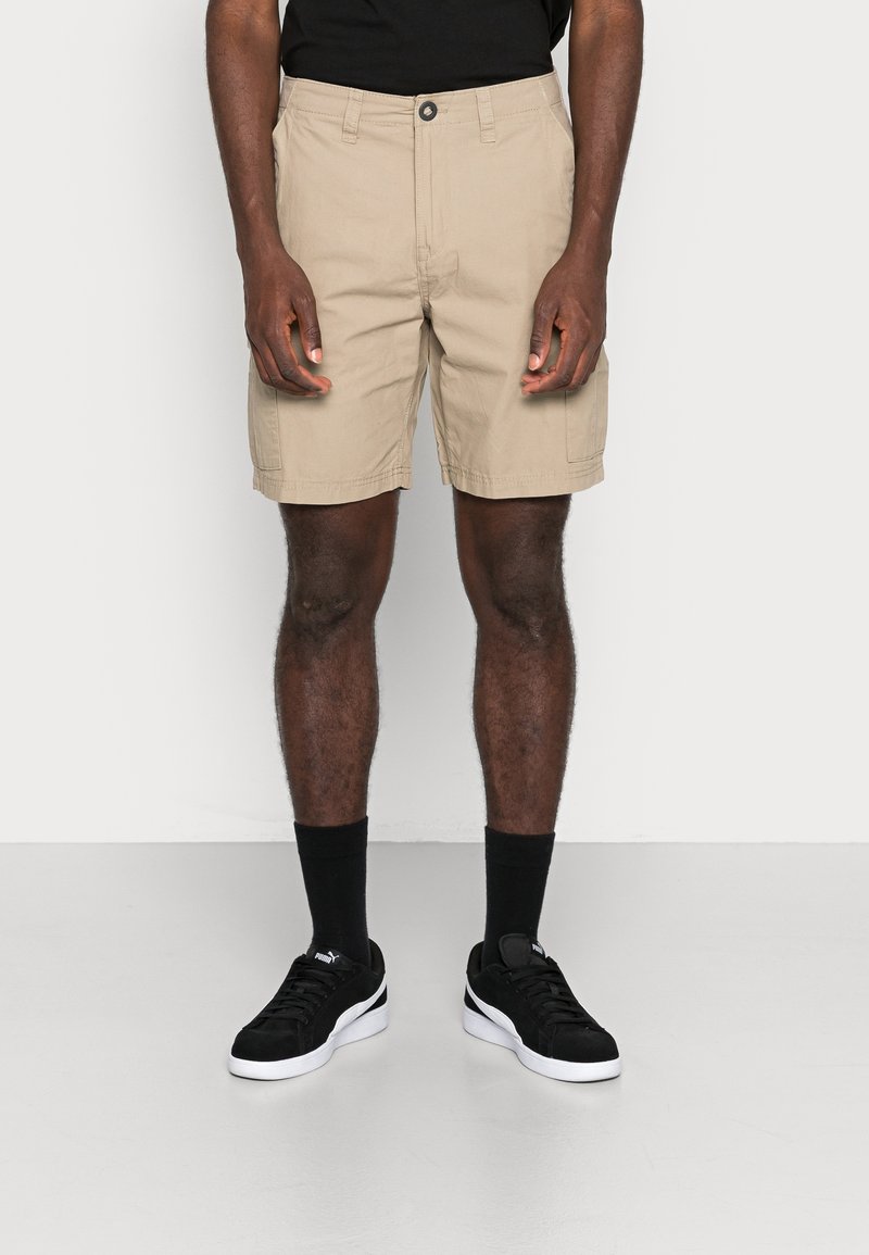 Homme portant un short cargo beige, des chaussettes noires et des baskets Puma noires, se tenant sur un sol clair contre un mur blanc.