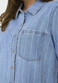 Ljust blå jeansskjorta med vertikala vita ränder, knappstängning, spetskrage och en enkel bröstficka. Mjuk textur och avslappnad design.