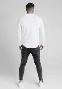 Vit långärmad skjorta med rundad nederkant, kombinerad med mörka, strukturerade tajta jeans och vita sneakers, sedd bakifrån.