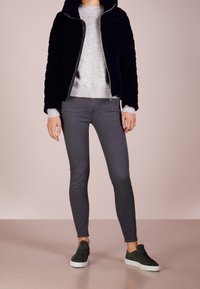 Marinblå quiltad jacka med huva, grå tröja, grå smala jeans och mörkgröna sneakers med vita sulor, presenterad mot en ljusrosa bakgrund.