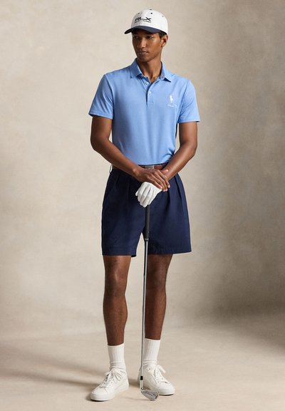 Uomo con polo azzurro chiaro, pantaloncini blu navy, guanto da golf bianco, cappellino bianco e sneakers che tiene una mazza da golf su sfondo semplice.