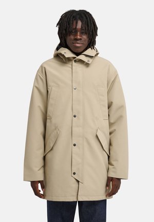 Jeune personne aux cheveux foncés portant un imperméable beige à capuche avec des poches avant et des boutons sur un pantalon foncé.