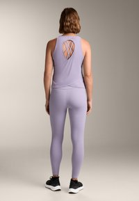 Fialový sportovní tank top s výřezem na zádech, v kombinaci s odpovídajícími legínami a černými teniskami. Hladká textilie.