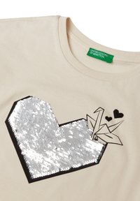 T-shirt in cotone beige con un grande design a cuore di paillettes argentate, contornato da nero, accompagnato da una gru origami nera e piccoli cuori.