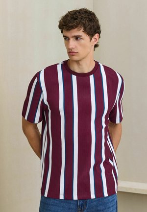 Giovane uomo con i capelli ricci che indossa una maglietta bordeaux a coste con righe verticali bianche e blu, che guarda di lato su uno sfondo semplice.
