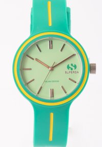Reloj de silicona verde con un anillo de acento amarillo, esfera minimalista verde menta, manecillas plateadas y logo que dice "SUPERGA". Presenta una textura suave.