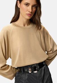 Sweat-shirt beige à manches longues avec un col rond, associé à un pantalon taille haute gris foncé et une ceinture noire avec des accessoires en métal argenté.