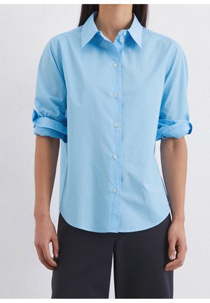 Persoon wearing een lichtblauw overhemd met opgerolde mouwen en zwarte broek, staand tegen een effen witte achtergrond.